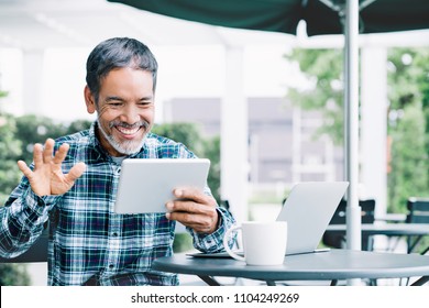 Mature Asian Man Waving Hand With Smiling And Enjoy Using Tablet Making Video Call To Child, Grandchild Or Friends At Outdoor Coffee Shop. Elderly People With Smart Technology Meeting Online Concept.