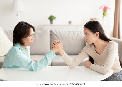 Mature Asian mother and adult daughter arm wrestling, looking each other in eyes, fighting at home. Parent child disagreement, confrontation between mother-in-law and daughter-in-law concept