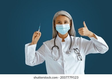 Mature Asian Woman Doctor In White Coat With Medical Mask Holds Syringe And Shows Thumb Up Standing On Blue Background In Studio. Vaccination Day