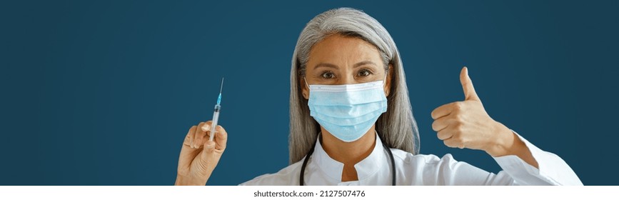 Mature Asian Woman Doctor In White Coat With Medical Mask Holds Syringe And Shows Thumb Up Standing On Blue Background In Studio. Vaccination Day