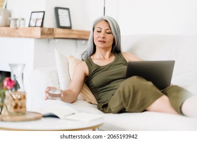 Mature Asian Woman Drinking Water While Working With Laptop On Couch At Home