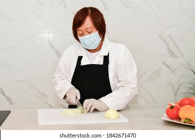 Mature Asian Woman With A Face Mask And Gloves Slicing A Peeled Onion On A Cutting Board At A Kitchen. Safety And Cooking Concept.