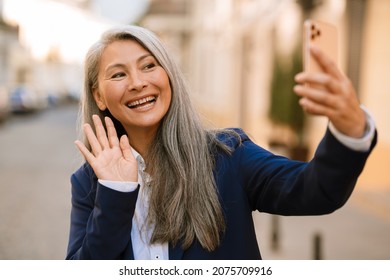 Mature Asian Woman Gesturing And Taking Selfie On Mobile Phone Outdoors