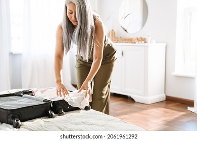 Mature asian woman with grey hair packing suitcase on bed at home
