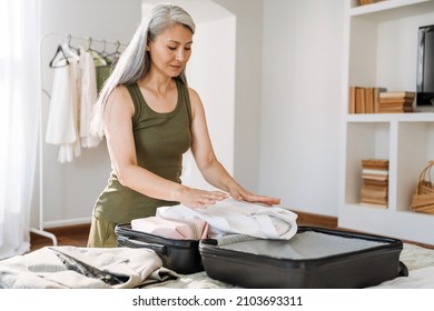 Mature Asian Woman With Grey Hair Packing Suitcase On Bed At Home