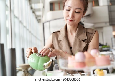 Mature Asian Woman Sitting At Table Eating Dessert Drinking Tea In Hotel Lobby Coffee Shop