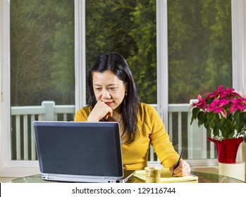 Mature Asian Woman Working At Home With Notebook Computer On Glass Table With Large Windows In Background