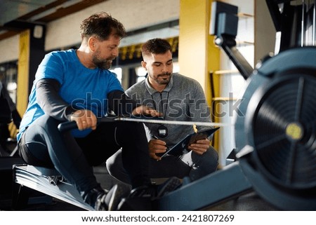 Mature athlete working out on rowing machine while analyzing exercise plan with his personal trainer in a gym.