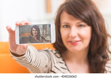 Mature Attractive Woman Taking Selfie Picture With Her White Smartphone At Home