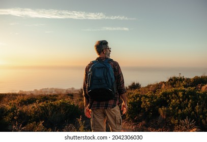 Mature backpacker looking at the panoramic view on a hilltop. Rearview of a male hiker standing alone on a coastal hill. Adventurous mature man enjoying the sunset outdoors.
