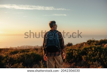 Mature backpacker looking at the panoramic view on a hilltop. Rearview of a male hiker standing alone on a coastal hill. Adventurous mature man enjoying the sunset outdoors.