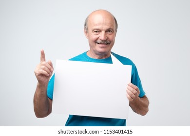 Mature Bald Man In Blue Tshirt Holding Blank Sheet Of Paper, Smiling. Isolated On White Wall.