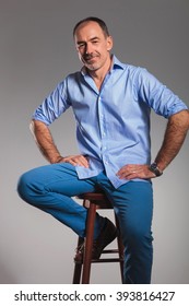 Mature Bearded Casual Man In Jeans Posing Seated With Hands On Waist Looking At The Camera In Gray Studio Background