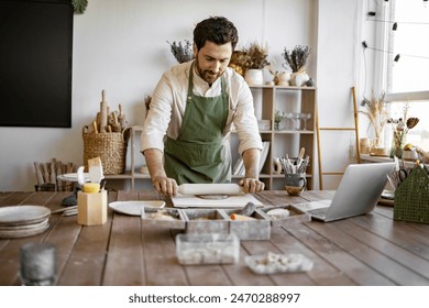 Mature bearded male potter flattens clay with rolling pin to create handmade dishes. Process of making piece of pottery art in bright studio against background of shelves with products. - Powered by Shutterstock