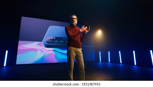 Mature Bearded Man In Casual Clothes Gesticulating And Talking About Innovative Features Of New Cellphone During Presentation On Stage Against LED Screen