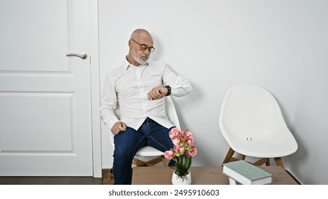 Mature bearded man in glasses checking time while sitting in a minimalist room with a white chair and door. - Powered by Shutterstock