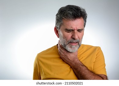 Mature Bearded Man Having Sore Throat And Touching His Neck, Wearing A Loose Yellow T-shirt Against Light Grey Background. Hard To Swallow. Terrible Pain In His Throat. 