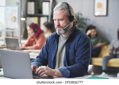 Mature Bearded Man In Headphones Working On Laptop At The Table In Call Center