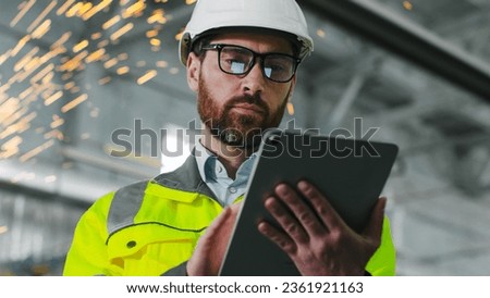 Mature bearded man on construction site. Engineer in uniform and glasses holds tablet background of workflow. An employee notes something in his gadget in workshop. Diversity of proffesions.