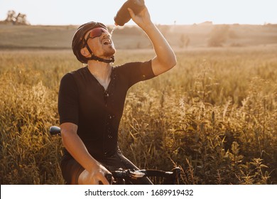 Mature bearded man in sport clothing sitting on his bike and drinking water from black bottle. Active cyclist  in helmet and eyewear feeling thirsty after long race.