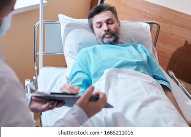 Mature Bearded Patient In Hospital Gown Lying In Bed And Signing Medical Agreement While Giving Consent To Surgical Operation