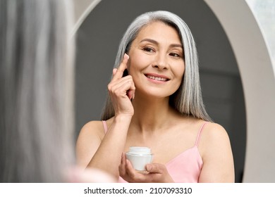 Mature Beautiful Happy Middle Aged Asian Woman, Older Grey Haired Lady Looking At Mirror Touching Face Applying Whitening Antiaging Anti Wrinkle Perfect Skin Care Cream.