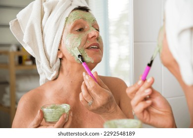 Mature beautiful woman applying green homemade DIY facial mask on skin. Looking in the mirror in bathroom, Wrapped in a towel. Skincare routine and anti-aging treatment at home for older women. - Powered by Shutterstock