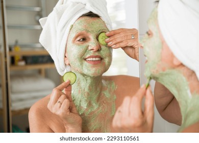 Mature beautiful woman applying green homemade DIY facial mask on skin. Looking in the mirror in bathroom, having fun. Wrapped in a towel. Skincare routine and anti-aging treatment for older women. - Powered by Shutterstock