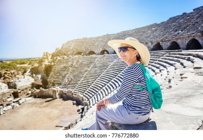 Mature Beautiful Woman Traveler, Sits On The Steps Of The Amphitheater In Admiring The View. Travel To Greece And Turkey, The Monuments Of Ancient Architecture, Active Seniors