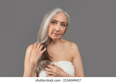 Mature Beauty. Feminine Senior Lady With Perfect Smooth Skin Wearing Towel After Bath, Posing To Camera Over Grey Studio Background. Portrait Of Older Woman Touching Her Long Hair