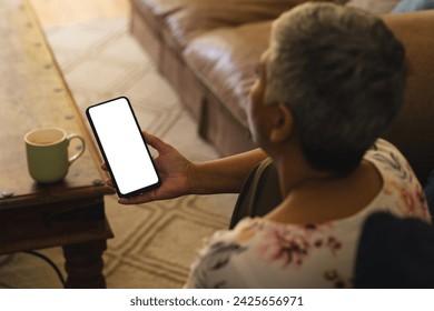 A mature biracial woman checks her phone at home, with copy space unaltered. She enjoys a quiet moment with a cup of coffee nearby. - Powered by Shutterstock