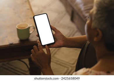 A mature biracial woman checks her smartphone at home, with copy space unaltered. She enjoys a quiet moment with a cup of coffee nearby. - Powered by Shutterstock