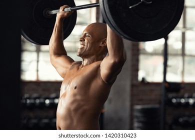 Mature Black Bodybuilder Doing Weight Lifting At Gym. Muscular African American Guy Doing Heavy Deadlift Exercise At Gym. Determined Strong Man Doing Exercises At Biceps And Shoulders.