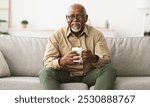 Mature Black Man Drinking Coffee Watching TV Sitting On Sofa And Smiling To Camera Holding Cup At Home, Wearing Eyeglasses. Retirement Lifestyle And Leisure Concept