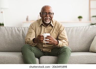 Mature Black Man Drinking Coffee Watching TV Sitting On Sofa And Smiling To Camera Holding Cup At Home, Wearing Eyeglasses. Retirement Lifestyle And Leisure Concept