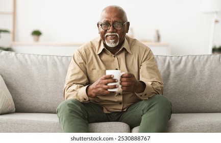 Mature Black Man Drinking Coffee Watching TV Sitting On Sofa And Smiling To Camera Holding Cup At Home, Wearing Eyeglasses. Retirement Lifestyle And Leisure Concept - Powered by Shutterstock