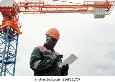 Mature Black Man In Gloves, Workwear And Hardhat Scrolling Through Online Information While Using Tablet At Construction Site