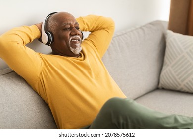 Mature Black Man Listening To Music Wearing Wireless Headphones And Holding Hands Behind Head Sitting On Couch At Home. Senior Male Relaxing Resting On Sofa Enjoying Favorite Song