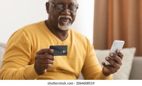 Mature Black Man Shopping Online Using Smartphone And Credit Card Making Payment Via Banking App Sitting On Sofa At Home. Selective Focus On Phone And Bank Card. Cropped, Panorama