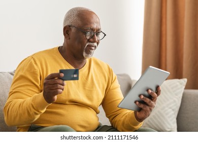 Mature Black Man Shopping Using Digital Tablet And Credit Card Making Payment Online Sitting On Sofa Indoors. Shopaholism, E-Commerce And Internet Banking Concept