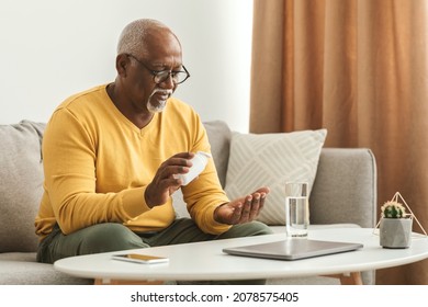 Mature Black Man Taking Medication Pill Putting Medicine On Hand Sitting Near Table With Laptop And Glass Of Water At Home. Medical Treatment, Supplements And Vitamins In Senior Age