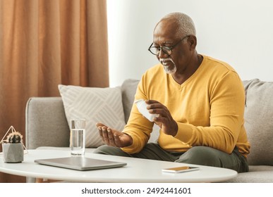 Mature Black Man Taking Medication Pill Putting Medicine On Hand Sitting Near Table With Laptop And Glass Of Water At Home. Medical Treatment, Supplements And Vitamins In Senior Age - Powered by Shutterstock