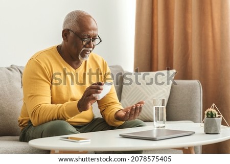 Mature Black Man Taking Medication Pill Putting Medicine On Hand Sitting Near Table With Laptop And Glass Of Water At Home. Medical Treatment, Supplements And Vitamins In Senior Age