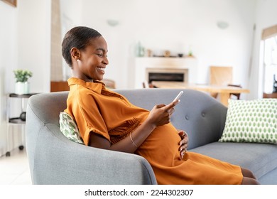 Mature black pregnant woman texting on mobile phone while sitting on couch in living room. Happy mid adult african american pregnant woman using smartphone while relaxing on sofa at home: motherhood. - Powered by Shutterstock