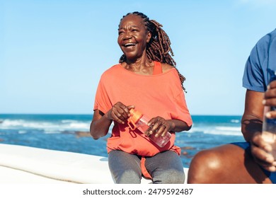 Mature black woman enjoying a sunny day by the seaside, smiling and holding a water bottle. She appears happy and relaxed. - Powered by Shutterstock