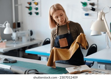 Mature blond craftswoman looking at piece of yellow leather or suede in her hands while choosing textile for new models of fashion collection - Powered by Shutterstock