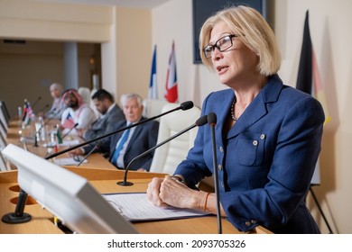 Mature blond female delegate in formalwear speaking in microphone while standing by tribune at political forum - Shutterstock ID 2093705326