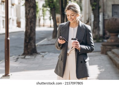 Mature blond smiling business woman walking around the city, drinking coffee and talking on the phone in spring on a background of lilac flowers