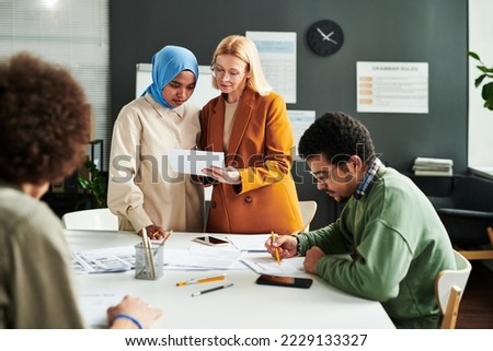 Mature blond teacher showing document with English grammar test to Muslim female student in hijab while both standing by workplace