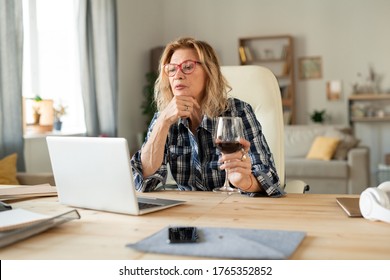 Mature blond woman in checkered shirt having glass of red wine while sitting by table in front of laptop and communicating online - Powered by Shutterstock